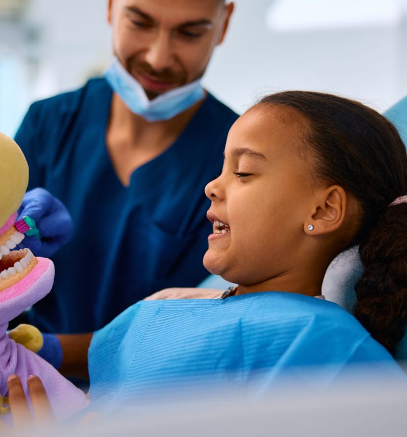 Dentist teaching African American little girl to brush teeth during appointment at dental clinic.